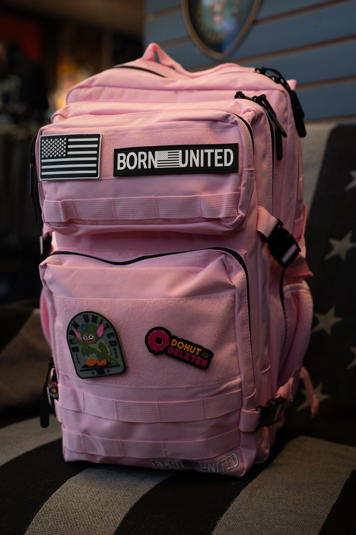 A pink tactical backpack with multiple zippered pockets, featuring a 'BORN UNITED' patch and other decorative patches, resting on a striped surface.