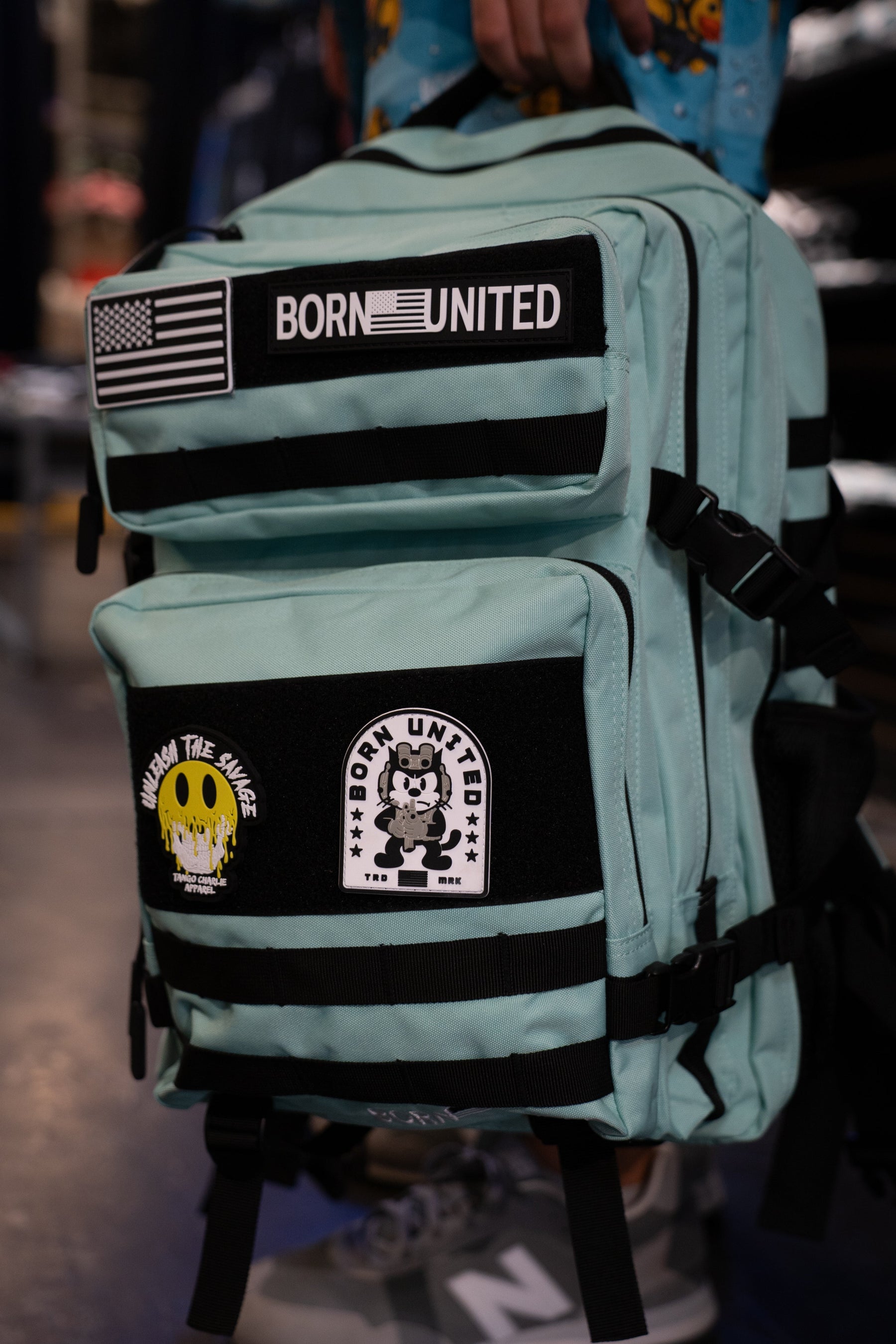 Introducing the Mint Condition Premium Tac-Pac - 45L by Born United Private Label: a mint green, TSA-approved travel backpack featuring black straps, an American flag patch, the BORN UNITED label, and two graphic patches—a bear and a smiley face.