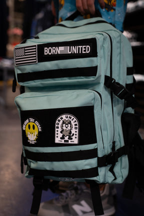 Introducing the Mint Condition Premium Tac-Pac - 45L by Born United Private Label: a mint green, TSA-approved travel backpack featuring black straps, an American flag patch, the BORN UNITED label, and two graphic patches—a bear and a smiley face.