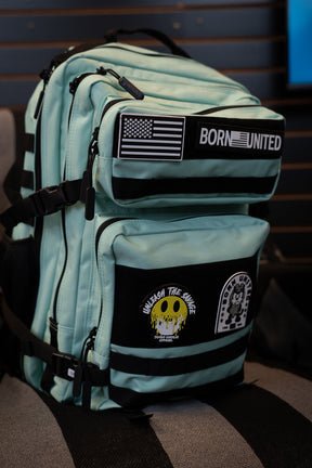 A Mint Condition Premium Tac-Pac - 45L by Born United Private Label, featuring a mint green and black design with multiple patches—including an American flag, "BORN UNITED," a skull graphic, and "UNLEASH THE SAVAGE"—rests on a striped chair.