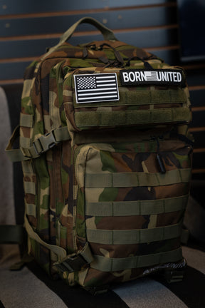 A tactical backpack in a classic woodland camouflage pattern, featuring a black and white American flag patch and a 'BORN UNITED' patch on the front flap, resting on a striped surface against a dark wood-paneled background.