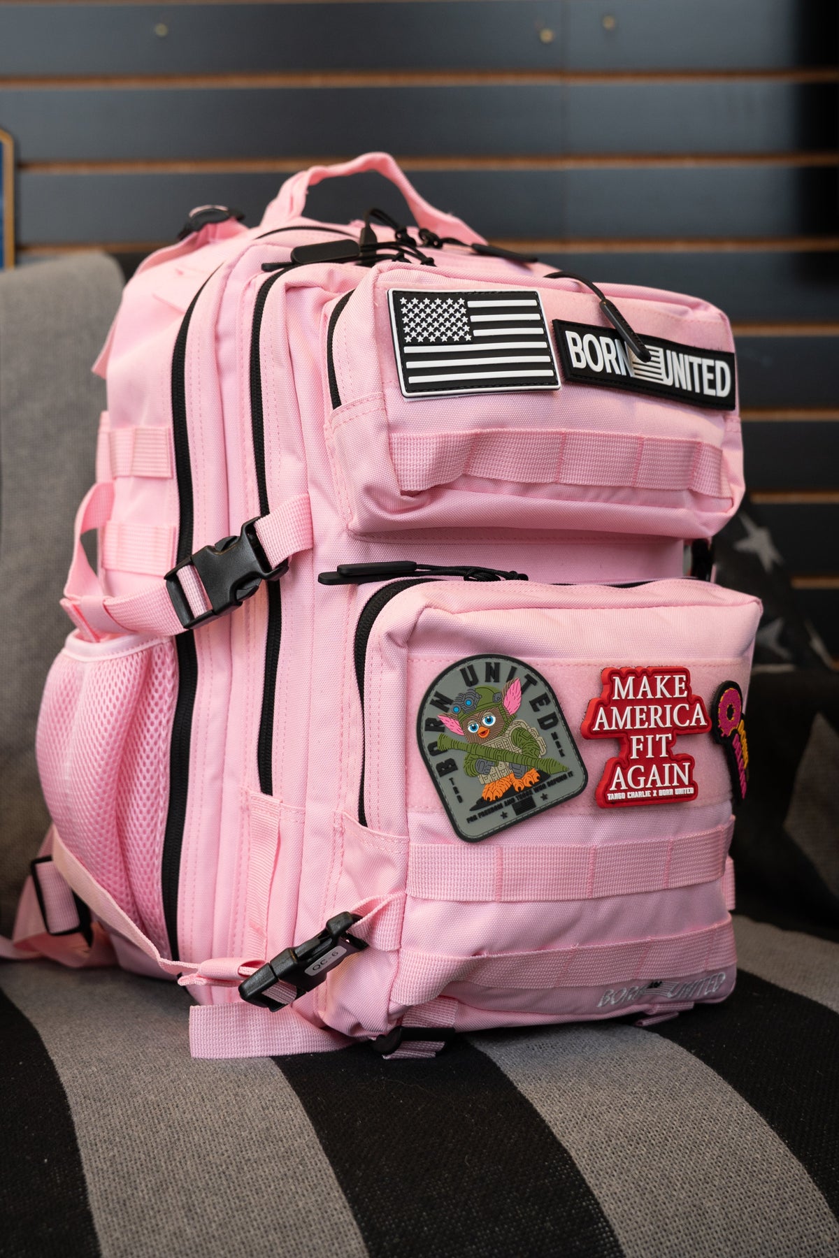A pink tactical backpack with black zippers and multiple patches, including a 'BORN TO LIMITED' patch and a 'MAKE AMERICA HIT AGAIN' patch, resting on a black and gray striped couch in front of a dark wood slat wall.