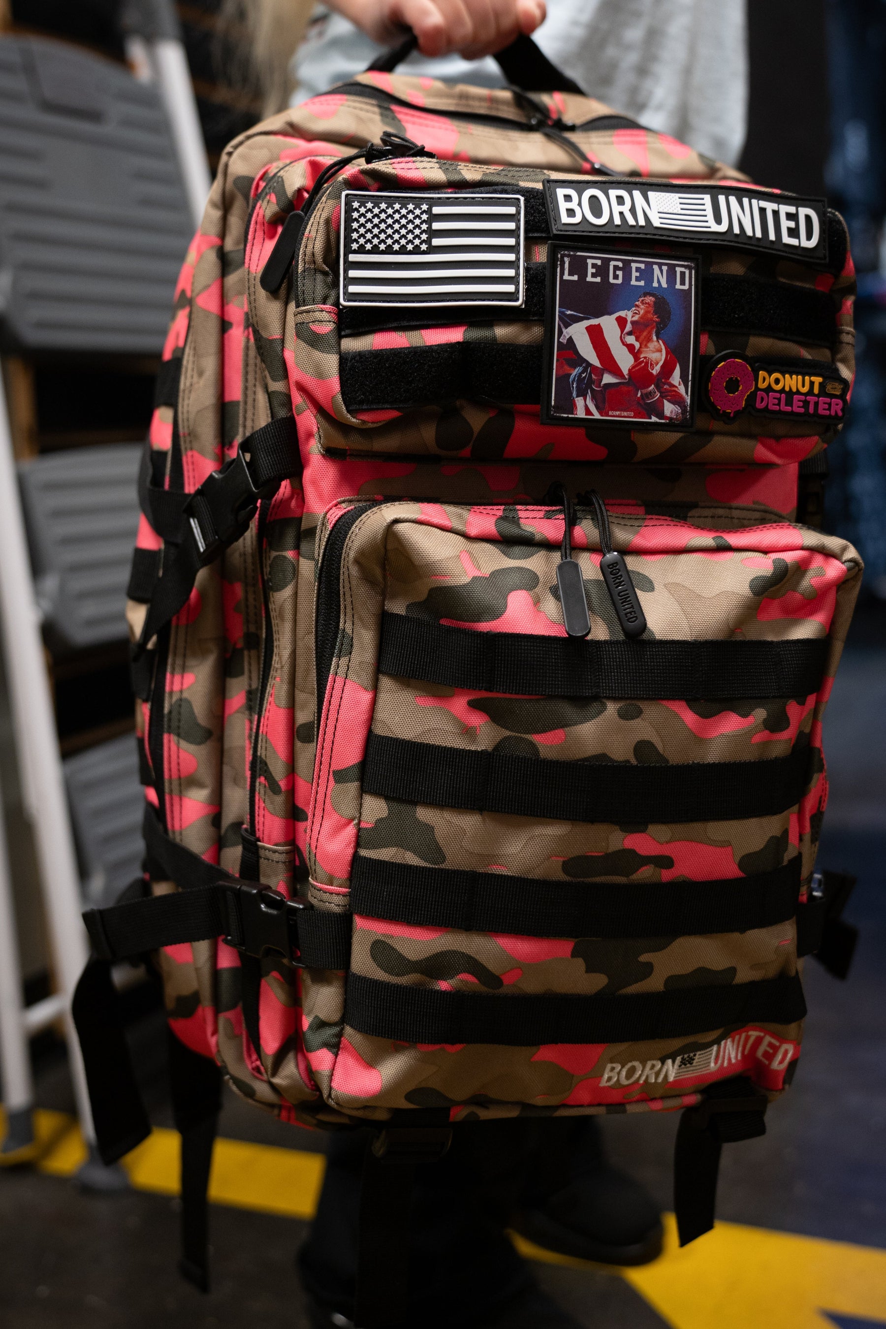 A person holds a Born United Private Label Multi-Cam Pink Tac-Pac - 45L backpack with pink accents, featuring patches including a U.S. flag, BORN UNITED, LEGEND, and DONUT DELETER.