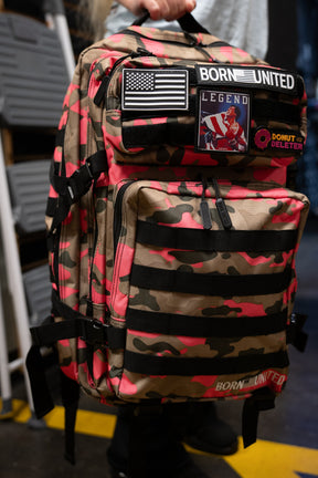 A person holds a Born United Private Label Multi-Cam Pink Tac-Pac - 45L backpack with pink accents, featuring patches including a U.S. flag, BORN UNITED, LEGEND, and DONUT DELETER.
