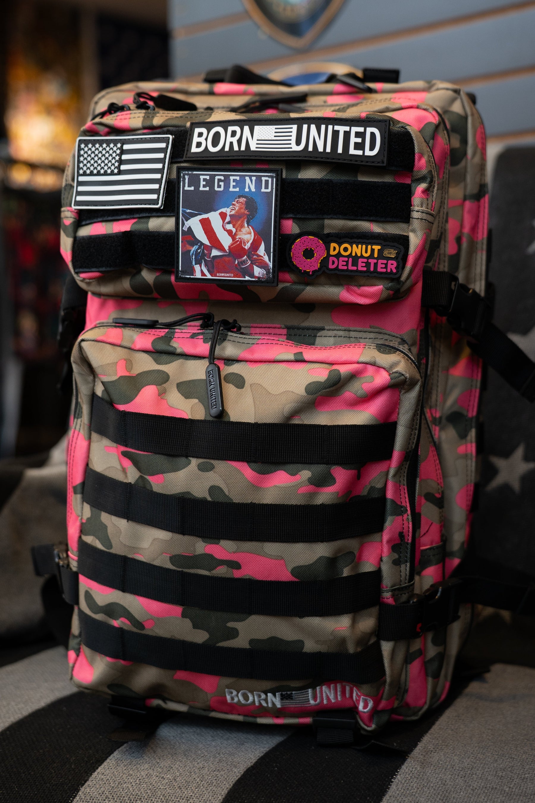 A pink and tan camouflage backpack with black webbing straps, multiple patches, and a 'BORN UNITED' logo, displayed on a black and gray striped cushion in a retail store.