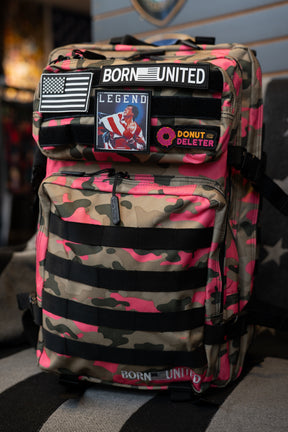 A pink and tan camouflage backpack with black webbing straps, multiple patches, and a 'BORN UNITED' logo, displayed on a black and gray striped cushion in a retail store.