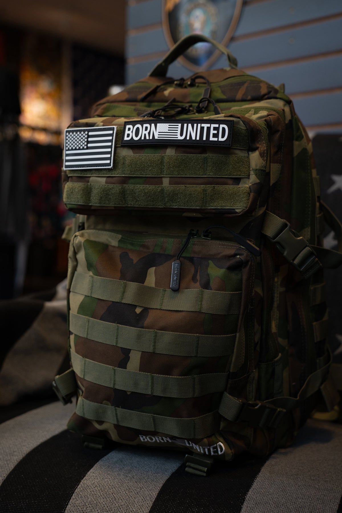 A Born United Private Label BDU Tac-Pac - 45L camouflage backpack, featuring velcro patches with "BORN UNITED" and an American flag, displayed indoors on a chair.