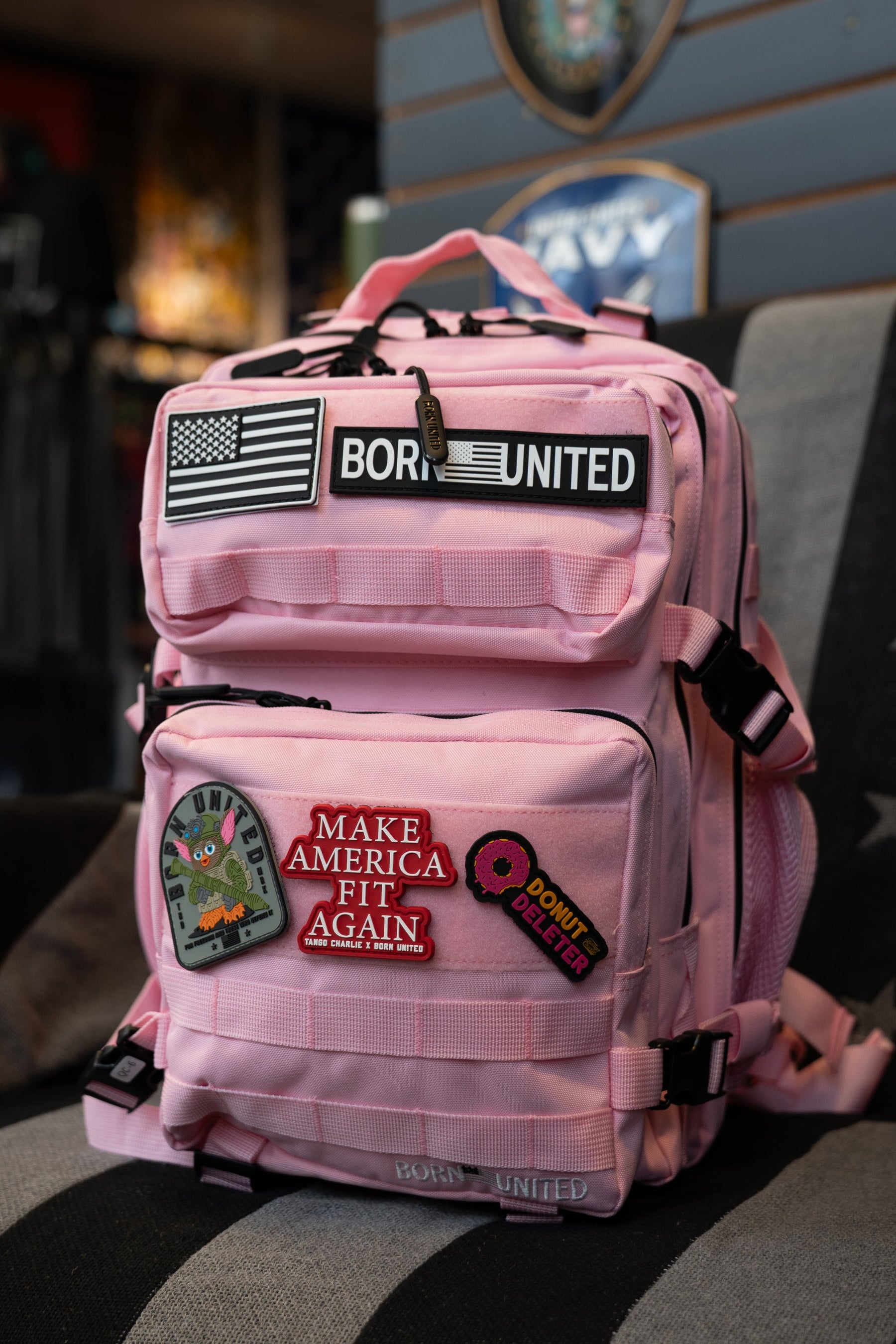 The Pink Mist Tac-Pac - 25L by Born United Private Label, featuring assorted patches such as an American flag, "Born United", "Make America Fit Again", a donut, and an alien, is displayed indoors on a chair.