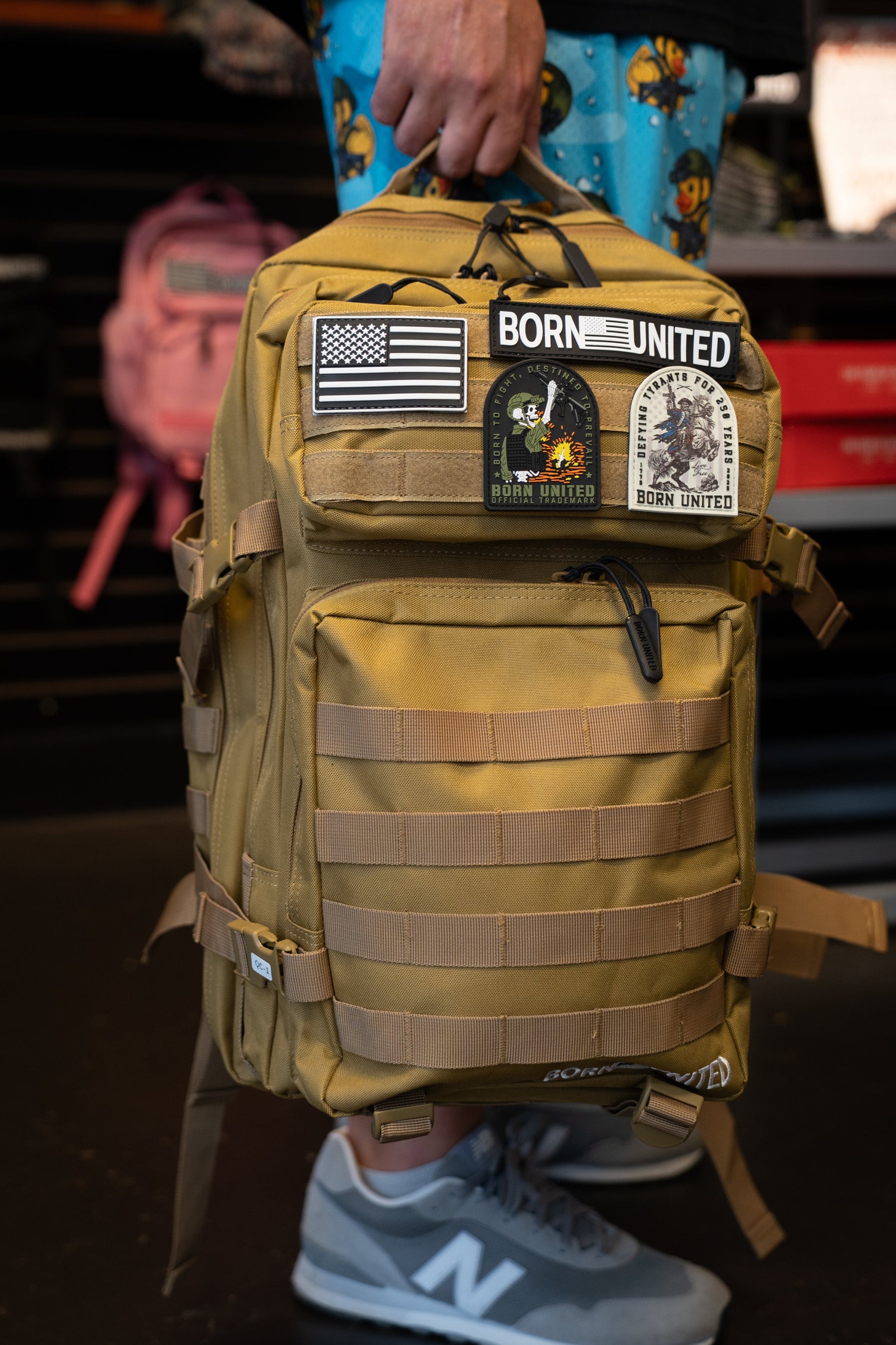 A person holds the Born United Private Label Coyote Brown Tac-Pac - 45L, wearing blue shorts with cartoon characters and gray New Balance sneakers.