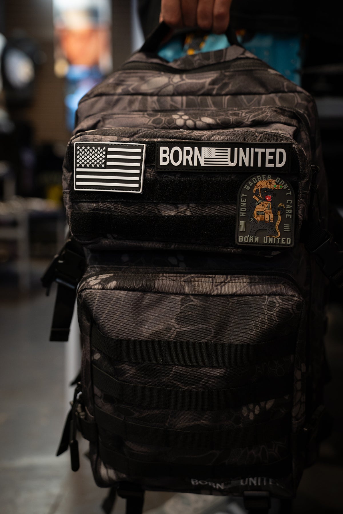 A person holds a black and gray camouflage backpack with 'BORN UNITED' patches, including an American flag patch, in a dimly lit indoor setting.