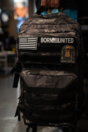 A person holds a black and gray camouflage backpack with 'BORN UNITED' patches, including an American flag patch, in a dimly lit indoor setting.