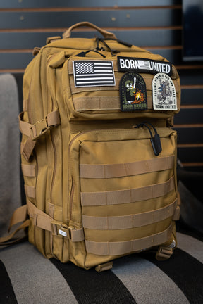 A Coyote Brown Tac-Pac - 45L tactical backpack from Born United Private Label, featuring multiple compartments and adorned with U.S. flag and “BORN UNITED” patches, sits on a gray and black striped chair.