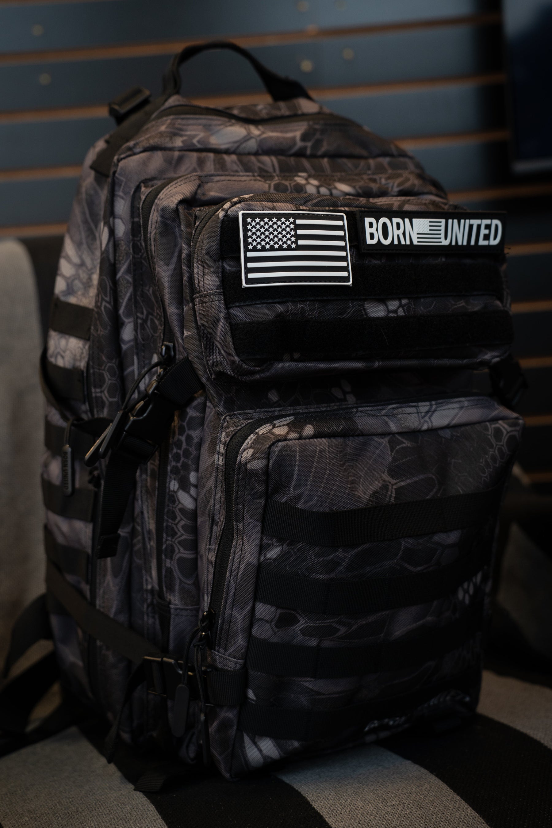 A black and gray camouflage backpack with a front pocket featuring a US flag patch and a 'BORN UNITED' logo patch, resting on a striped surface against a dark slatted wall.