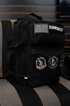 A black tactical backpack with multiple patches, including a US flag patch and 'BORN UNITED' text patch, resting on a gray and black striped couch in front of a dark wood slat wall.