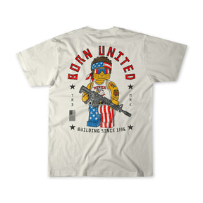 The Brick America white t-shirt by Born United Private Label showcases a muscular teddy bear in patriotic gear, wielding a rifle and shield with the US flag. "BORN UNITED" and "BUILDING SINCE 1776" text highlight its patriotic brick strength.