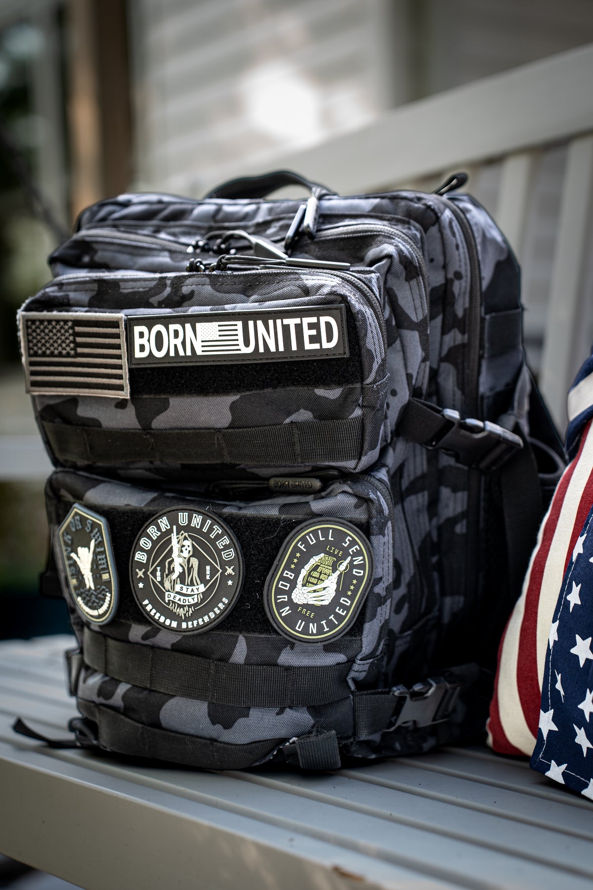 The Black Camo Tac-Pac - 25L by Born United Private Label, featuring multiple patches like a U.S. flag and BORN UNITED logo, rests on a bench next to a pillow with a stars and stripes motif.