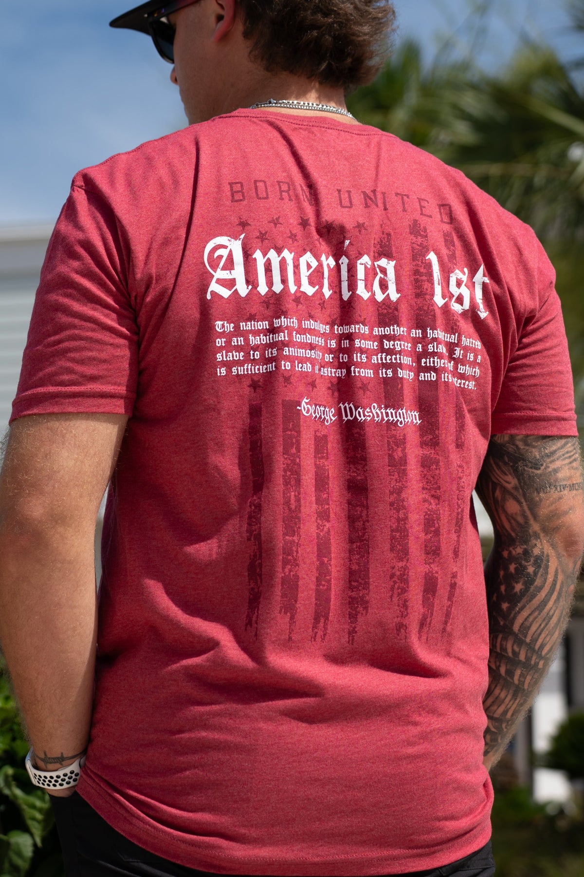 A man outdoors wears an America First t-shirt by Born United Private Label. The red shirt's back displays faded flag graphics and a George Washington quote in white text. He also sports sunglasses and a watch.