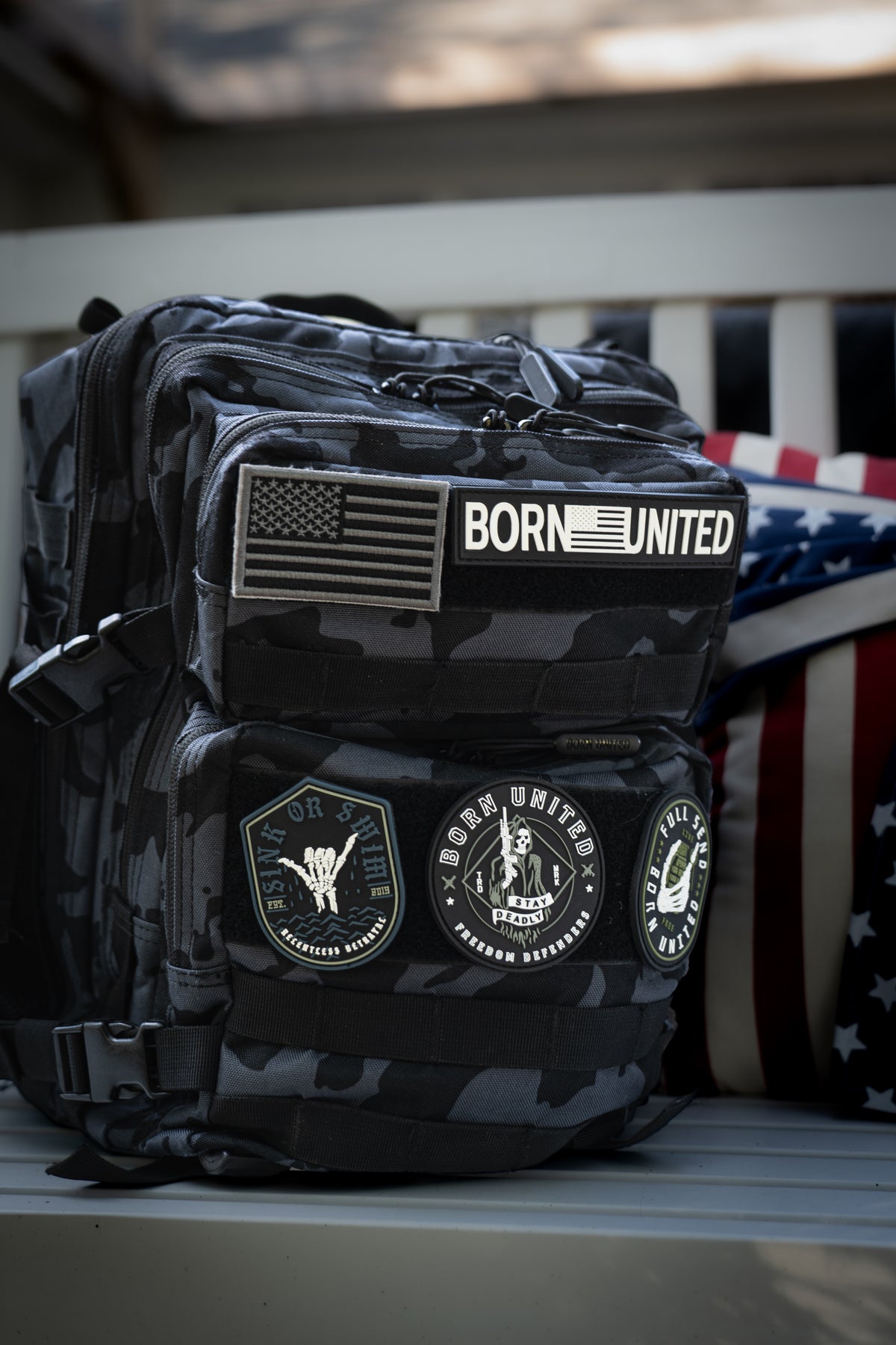 The Born United Private Label Black Camo Tac-Pac - 25L, featuring military-style patches like BORN UNITED and an American flag, rests on a bench beside a stars and stripes pillow.