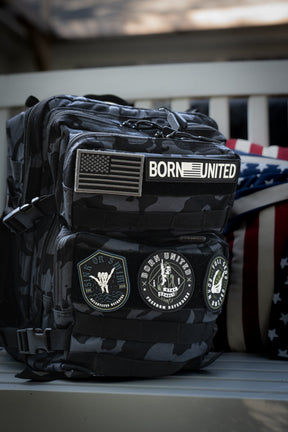 The Born United Private Label Black Camo Tac-Pac - 25L, featuring military-style patches like BORN UNITED and an American flag, rests on a bench beside a stars and stripes pillow.