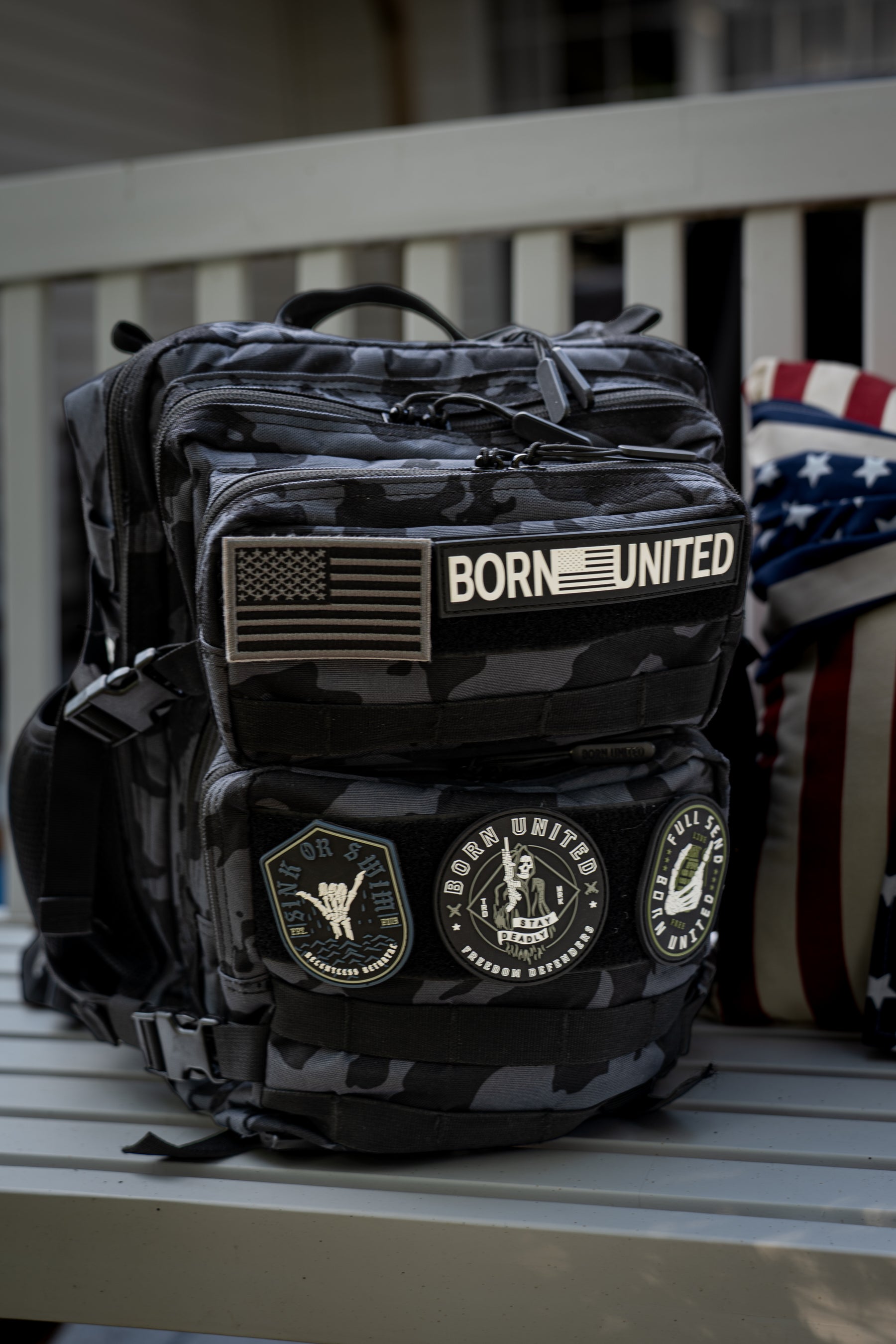 A Black Camo Tac-Pac - 25L by Born United Private Label sits on a bench, featuring an American flag patch, a BORN UNITED patch, and military-style round patches. A partially visible American flag pillow is behind the backpack.