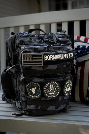 A Black Camo Tac-Pac - 25L by Born United Private Label sits on a bench, featuring an American flag patch, a BORN UNITED patch, and military-style round patches. A partially visible American flag pillow is behind the backpack.