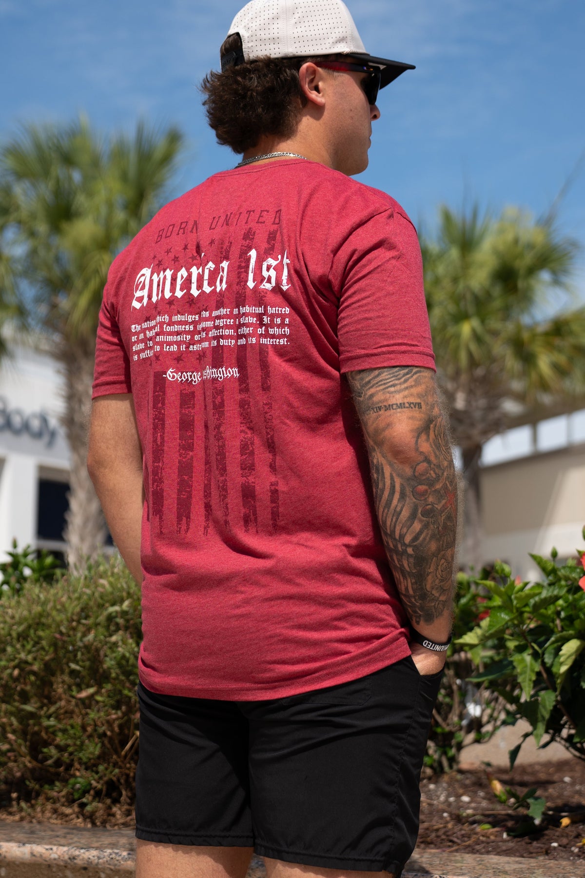 A man stands outdoors in black shorts, a white mesh cap, and sunglasses with palm trees and a building behind him. He wears a red "America First" t-shirt from Born United Private Label and has a large George Washington tattoo on his right arm.
