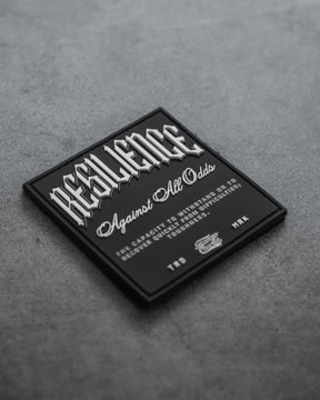The Resilience-PVC Patch by Tango Charlie Apparel features white text, RESILIENCE Against All Odds: The capacity to withstand or to recover quickly from difficulties; toughness, on a black square patch displayed on a gray textured surface.
