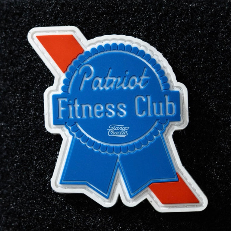 The Patriots Fitness Club - Patch by Tango Charlie Apparel is a blue badge-shaped 3D PVC patch with red accents and "Patriot Fitness Club" in white and blue text, shown against a black textured background.