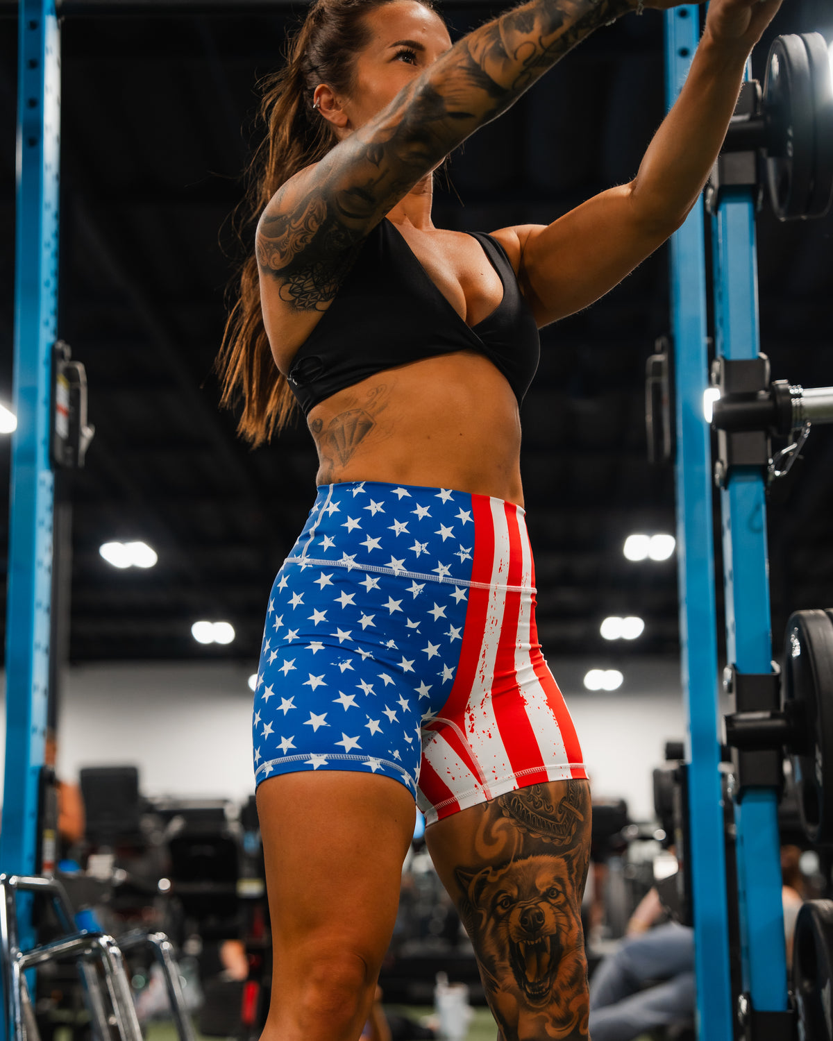 A woman with tattoos and long hair works out in a gym, wearing Born United Private Labels Womens High Waisted Seamless Front Compression Training Shorts | Stars & Stripes. Gym equipment is visible in the background.
