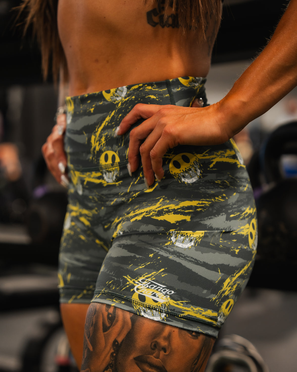 A person in Born United Private Labels Womens High-Waisted Seamless Front Compression Training Shorts, gray and yellow with skull designs, stands in the gym. Their hand is on their hip, showing part of a detailed thigh tattoo.