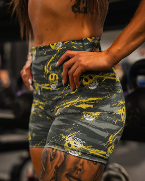 A person in Born United Private Labels Womens High-Waisted Seamless Front Compression Training Shorts, gray and yellow with skull designs, stands in the gym. Their hand is on their hip, showing part of a detailed thigh tattoo.