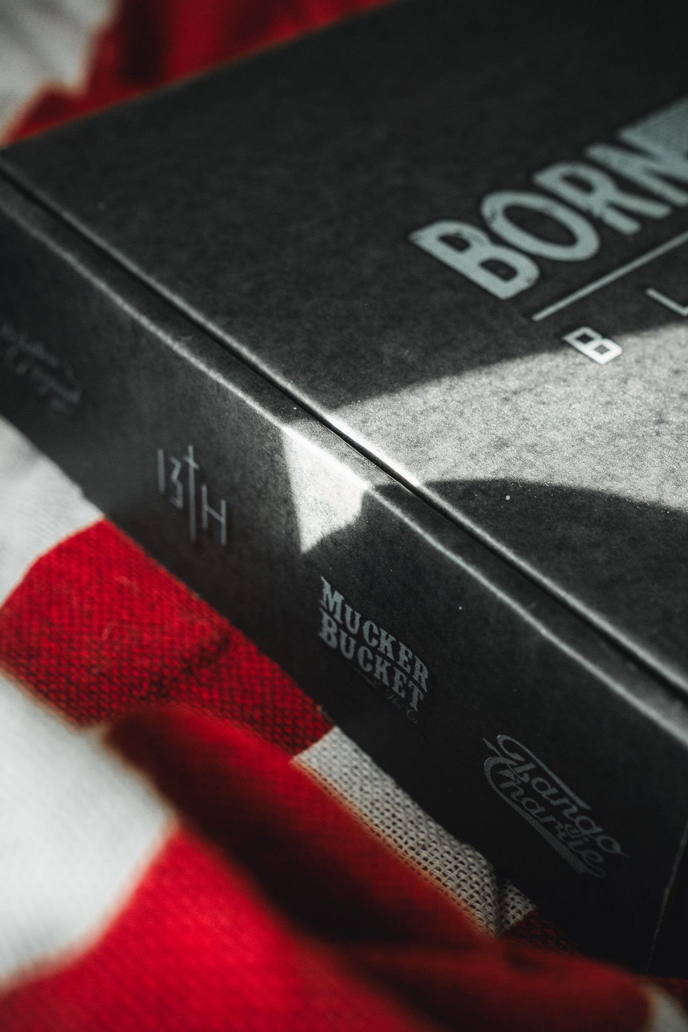 A close-up of a black Born United Black Box - Tactigator book by Born United Private Label rests on red-and-white striped fabric, with logos and text hinting at the intrigue of a mystery release.