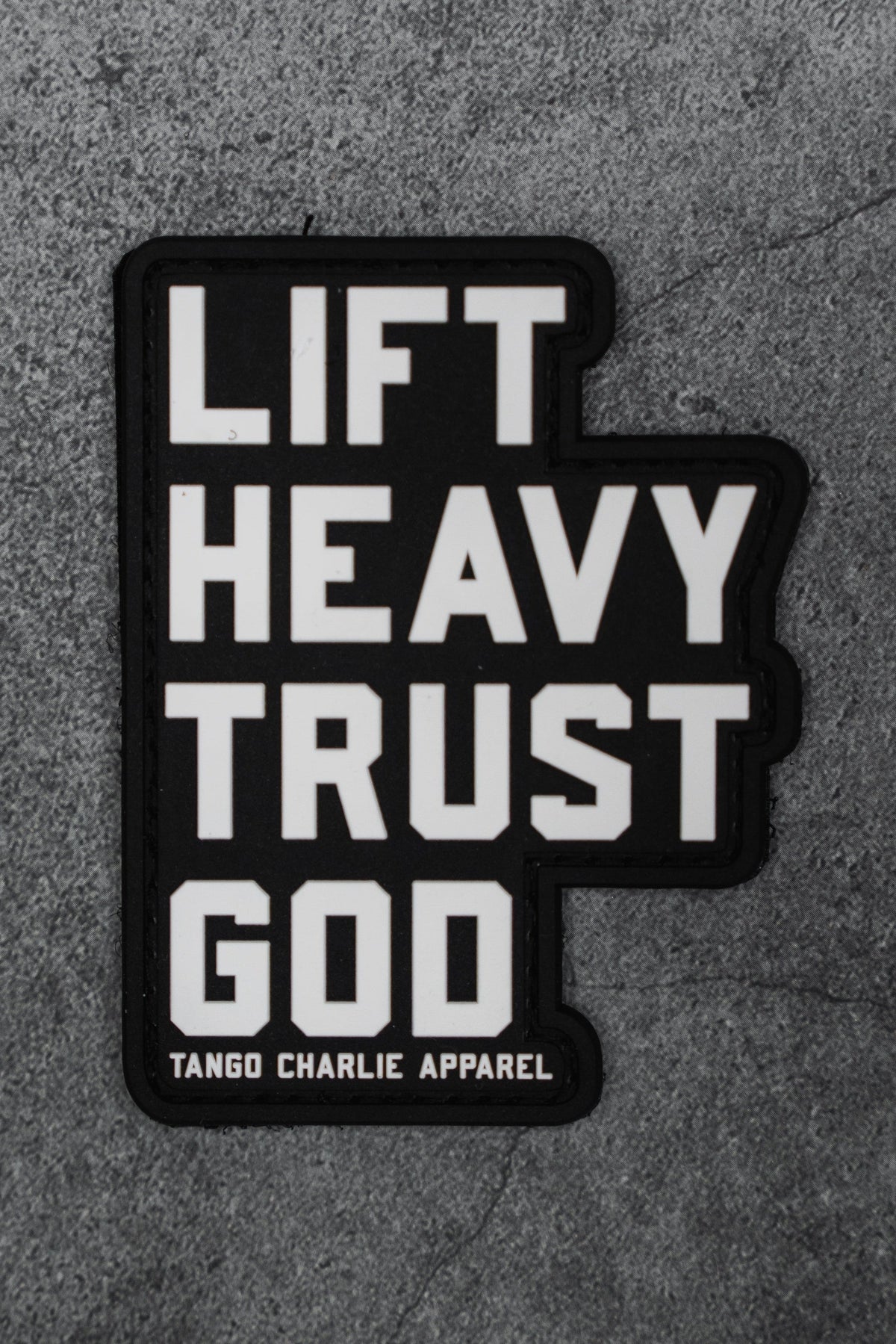 The Lift Heavy. Trust God. - Patch by Tango Charlie Apparel is a black 3D PVC patch with bold white text, shown on a gray textured background.