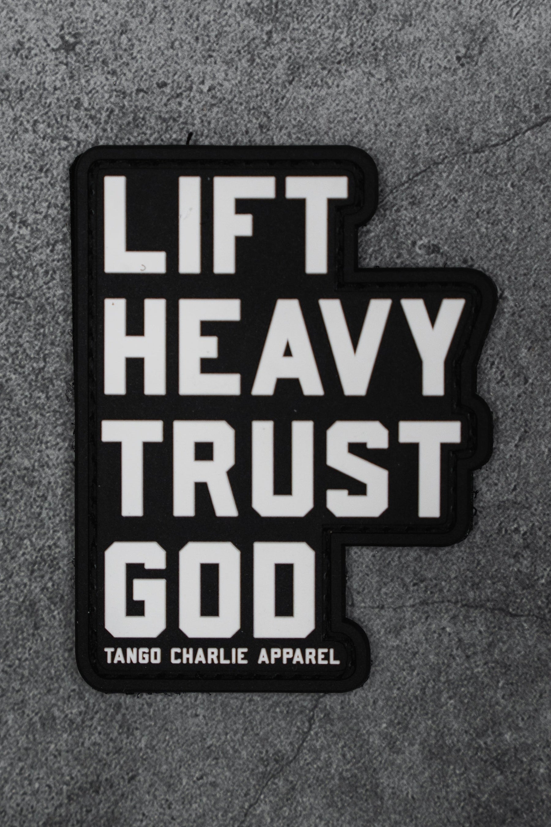 The Lift Heavy. Trust God. - Patch by Tango Charlie Apparel is a black 3D PVC patch with bold white text, shown on a gray textured background.