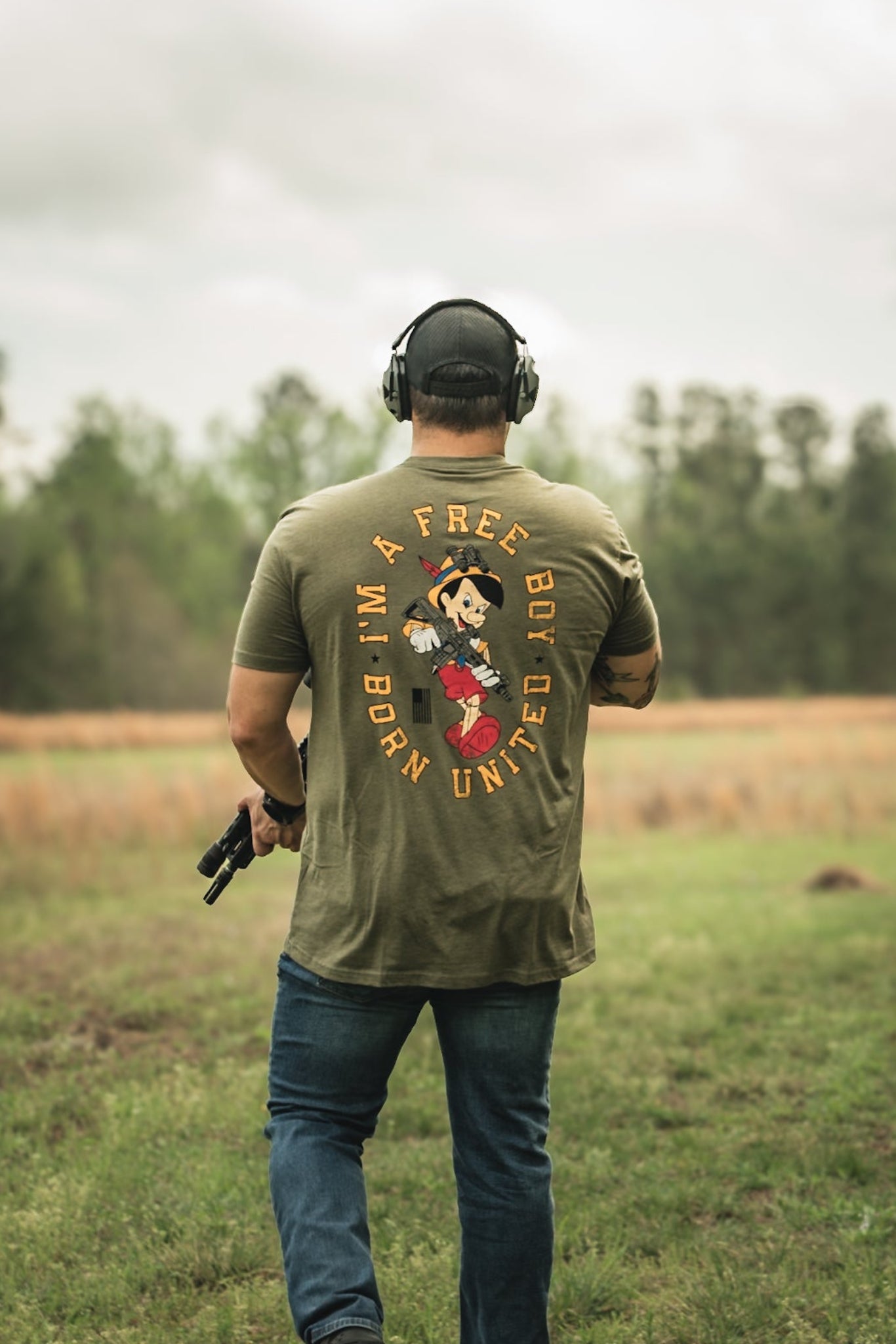 A boy in the Free Boy Tee by Born United Private Label, featuring headphones and a cartoon character on the back, stands with a handgun in a grassy field. Trees and clouds fill the background.