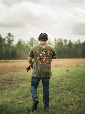 A boy in the Free Boy Tee by Born United Private Label, featuring headphones and a cartoon character on the back, stands with a handgun in a grassy field. Trees and clouds fill the background.