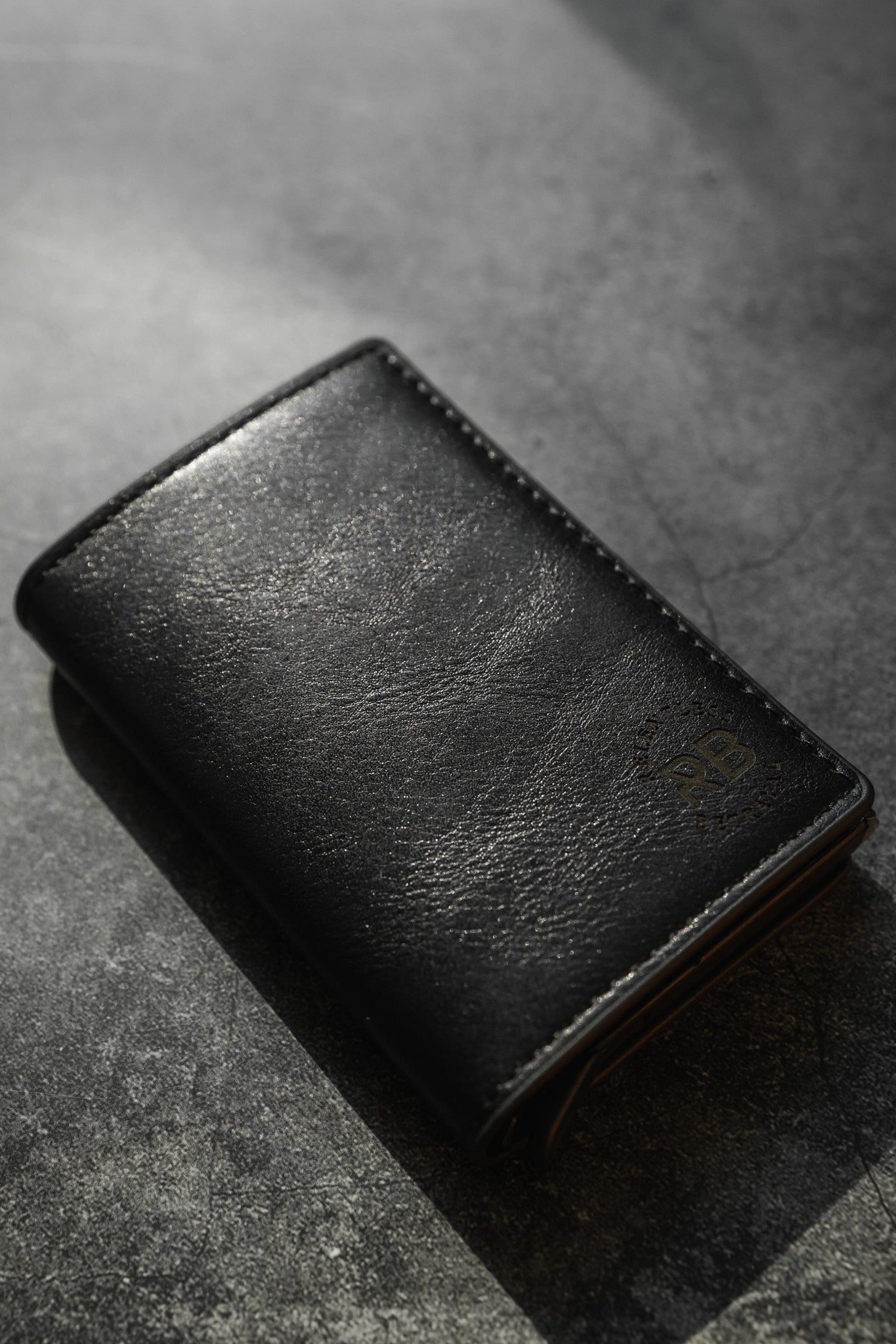 A Relentless Betrayal Minimalist Wallet in black leather rests on a textured dark gray surface. Soft lighting highlights its smooth texture, subtle stitching, and the embossed logo in the bottom right corner.