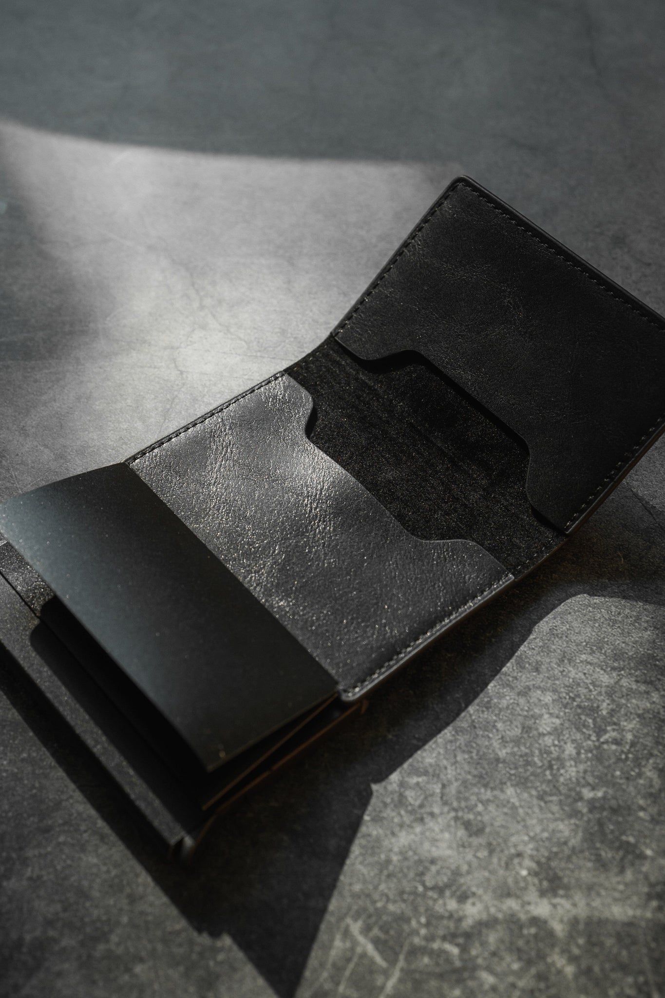 A black leather RFID-blocking Minimalist Wallet by Relentless Betrayal lies open on a textured gray surface, casting shadows. Its pop-up design reveals two visible compartments for credit card protection, yet it remains empty.