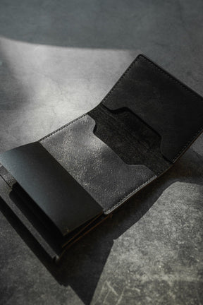 A black leather RFID-blocking Minimalist Wallet by Relentless Betrayal lies open on a textured gray surface, casting shadows. Its pop-up design reveals two visible compartments for credit card protection, yet it remains empty.