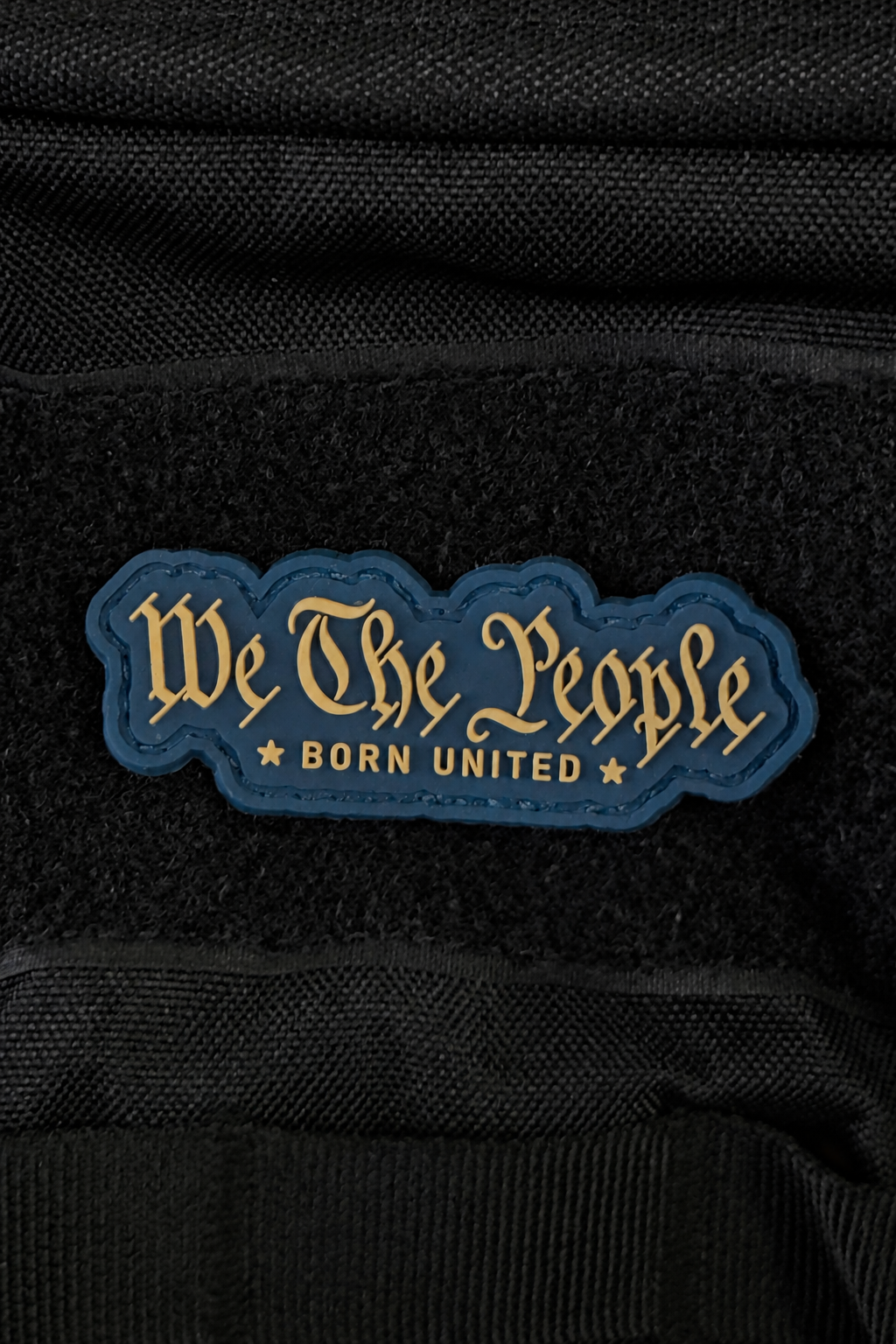 We The People PVC Patch in blue, 3 inches, featuring gold stylized text, BORN UNITED with two stars underneath, attached to a black textured surface.