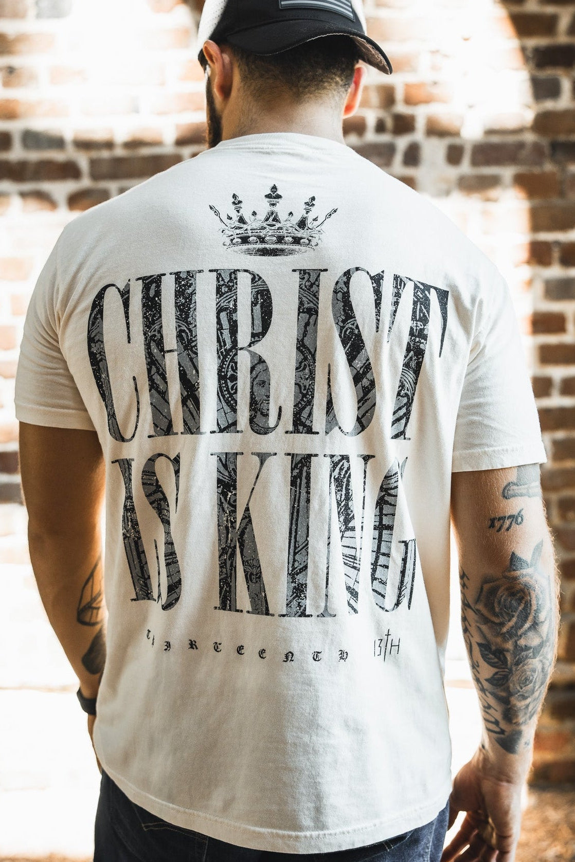 A man wearing the 13th Christ is King Premium Tee - Ivory, featuring "CHRIST IS KING" and a crown on the back, stands by a brick wall with tattoos on his right arm and an American flag patch on his cap.