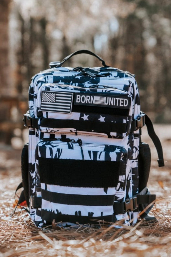 The Born United Private Label American Stealth Special Edition Tac-Pac - 25L, featuring a black-and-white U.S. flag design, MOLLE webbing, and a BORN UNITED patch, rests on dry pine needles with blurred trees behind.