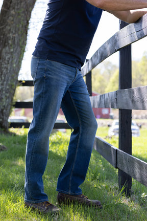 Wearing Devil Dog Dungarees Relaxed Straight Jean - Bryson City, a person in a navy shirt and brown shoes stands on grass by a wooden fence in sunlight.