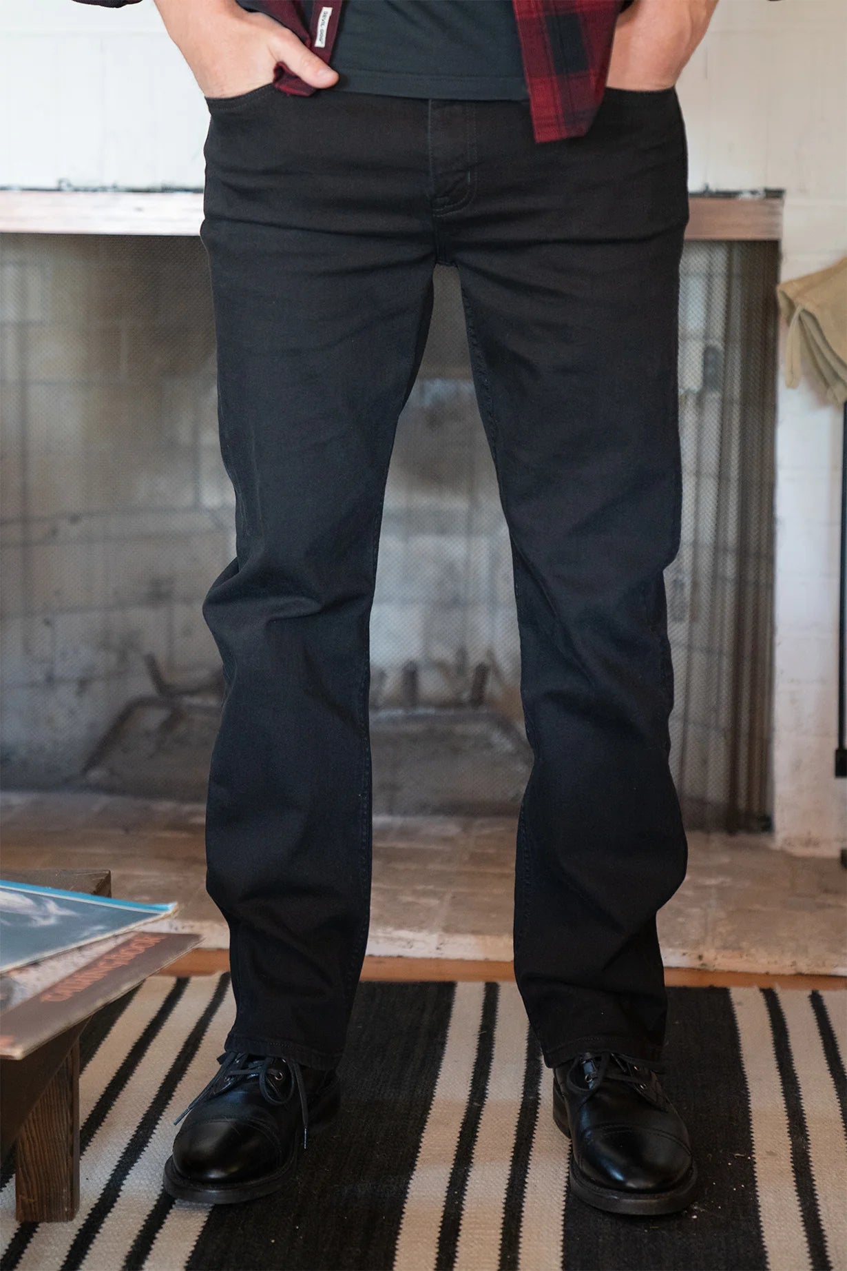 A person stands indoors on a black-and-white striped rug wearing Devil Dog Dungarees’ Relaxed Straight Jean - Cornelius in black, with black shoes and a red plaid shirt. A fireplace and table appear in the background.