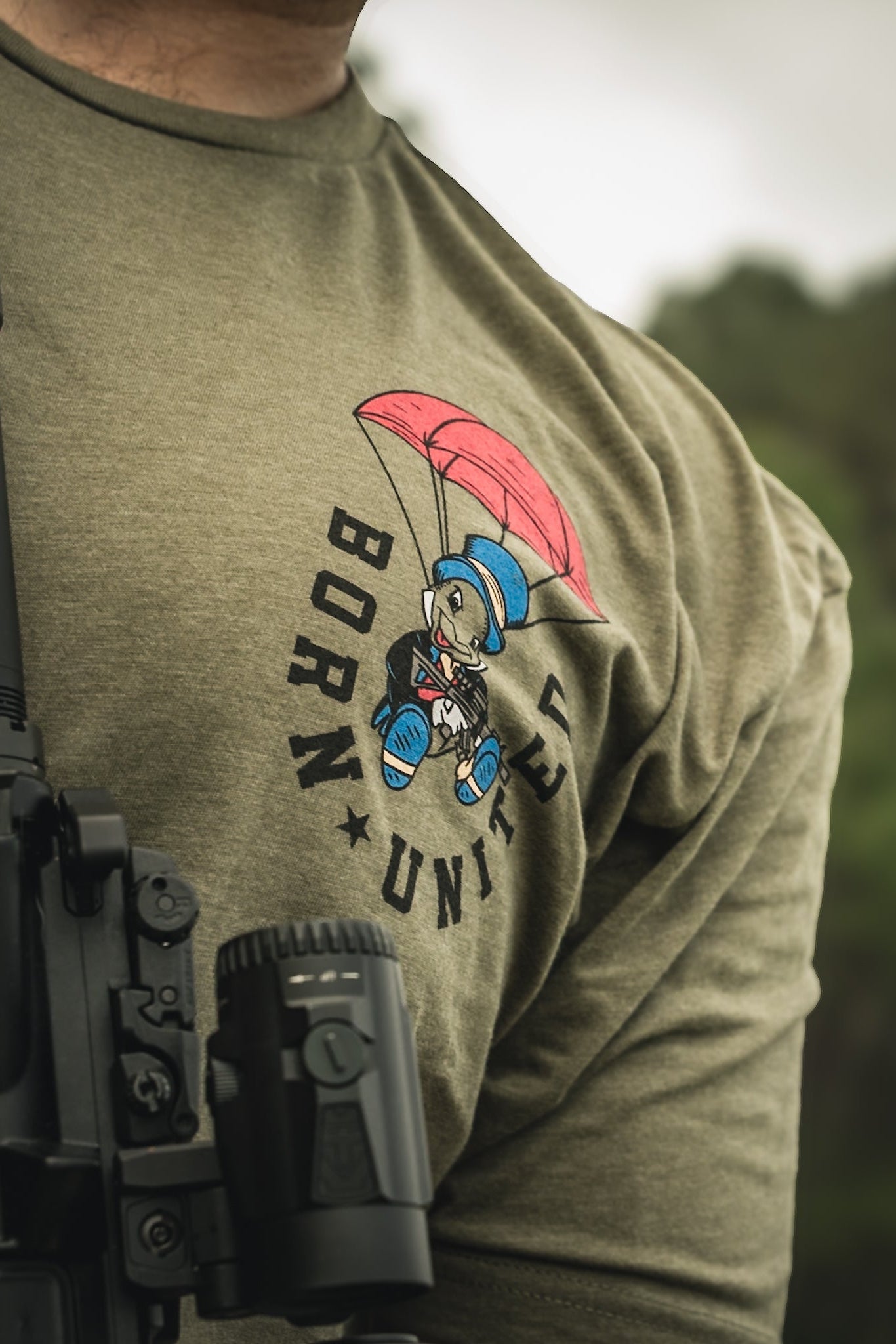 A person models the Free Boy Tee by Born United Private Label, featuring an olive green T-shirt with a cartoon bear in a blue helmet and red shoes parachuting. BORN UNITED is printed in black. A black machine is partly visible in front.