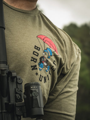 A person models the Free Boy Tee by Born United Private Label, featuring an olive green T-shirt with a cartoon bear in a blue helmet and red shoes parachuting. BORN UNITED is printed in black. A black machine is partly visible in front.