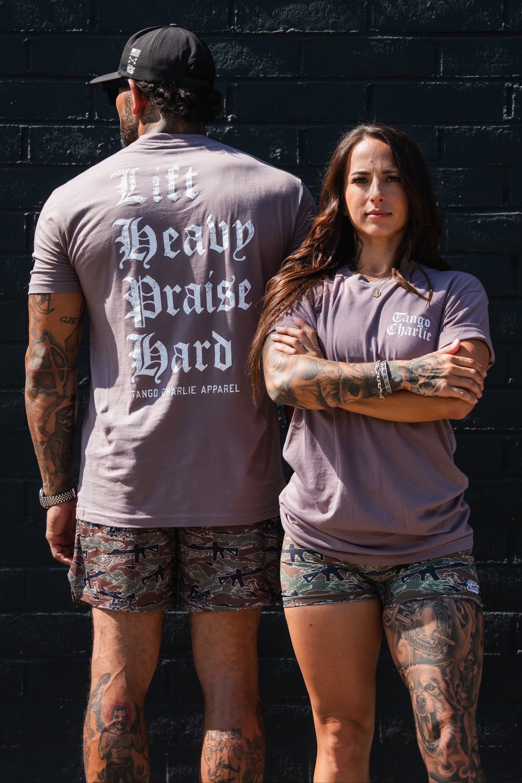 A man and woman stand back to back in matching mauve Lift Heavy. Praise Hard. Shiitake Tees and camouflage shorts; the man’s shirt displays the words in large Gothic lettering.