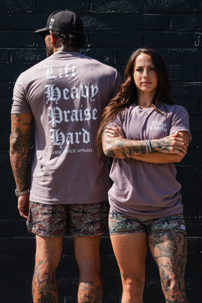 A man and woman stand back to back in matching mauve Lift Heavy. Praise Hard. Shiitake Tees and camouflage shorts; the man’s shirt displays the words in large Gothic lettering.
