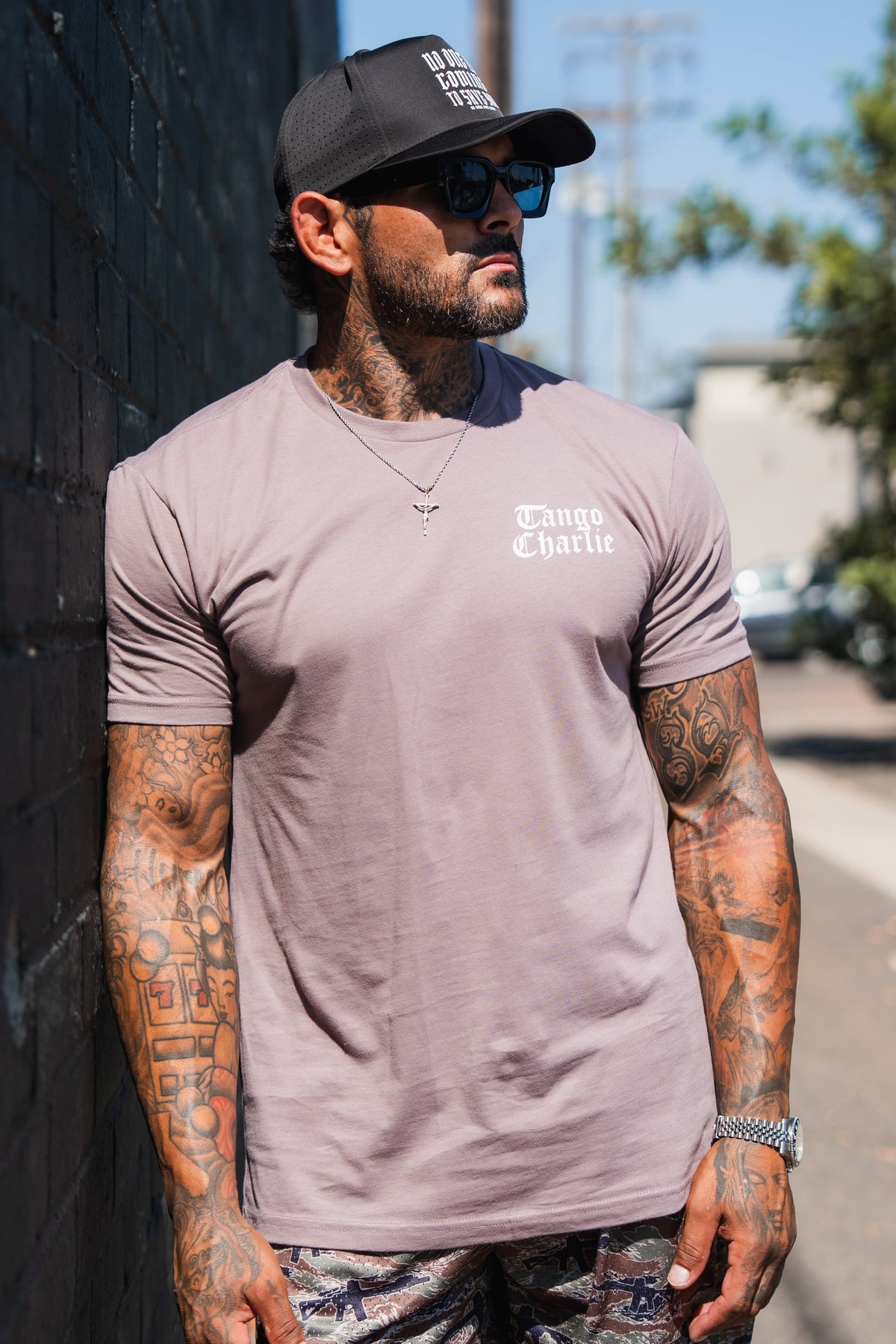 A man with tattoos on his arms and neck wearing a mauve Lift Heavy. Praise Hard. Shiitake Tee, sunglasses, and a black baseball cap.