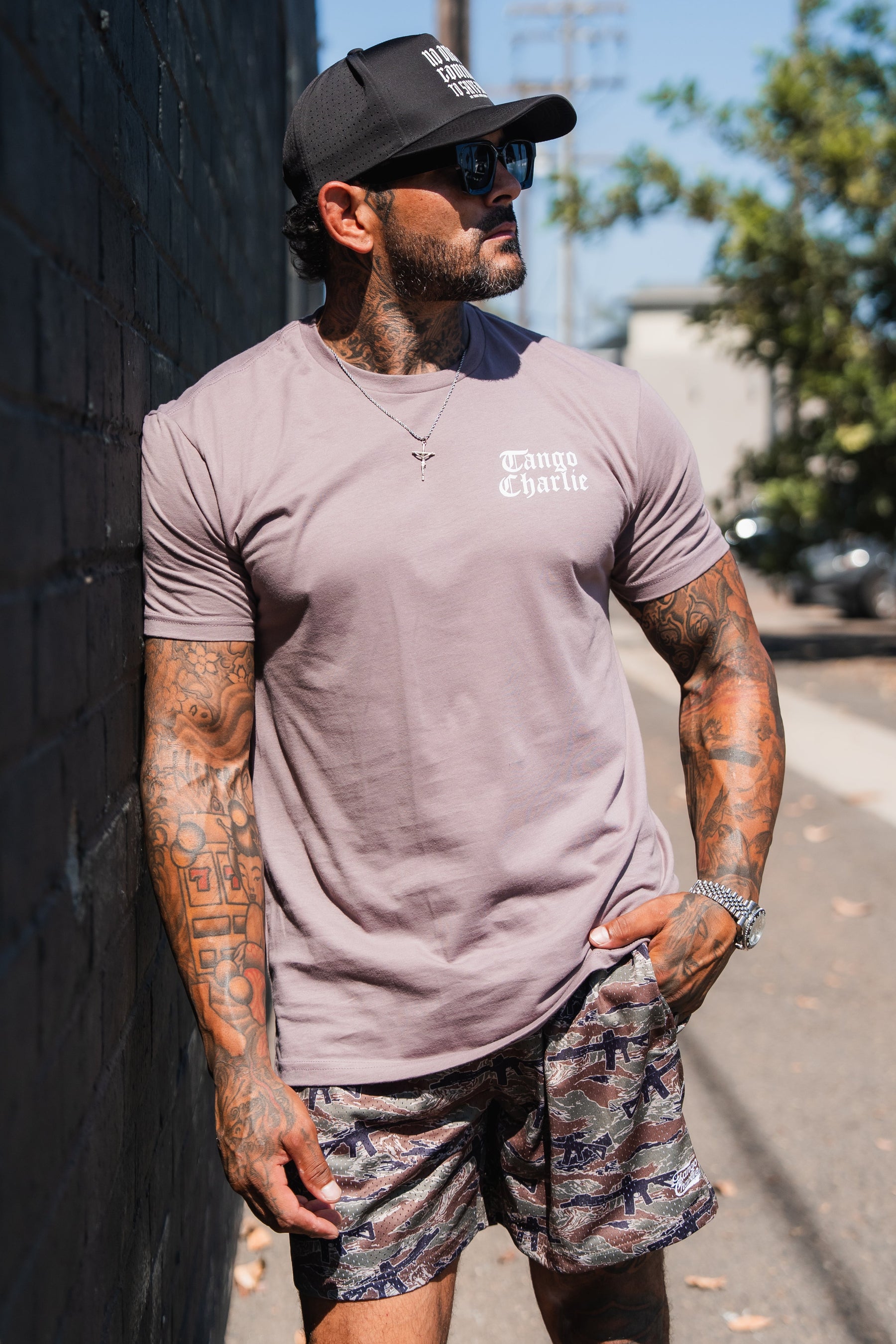 A man wearing a mauve Lift Heavy. Praise Hard. Shiitake Tee, a black baseball cap, and camouflage shorts.
